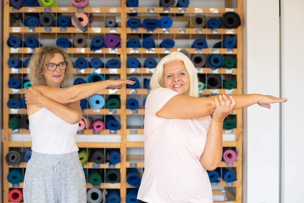 Two older women doing a standing arm stretch