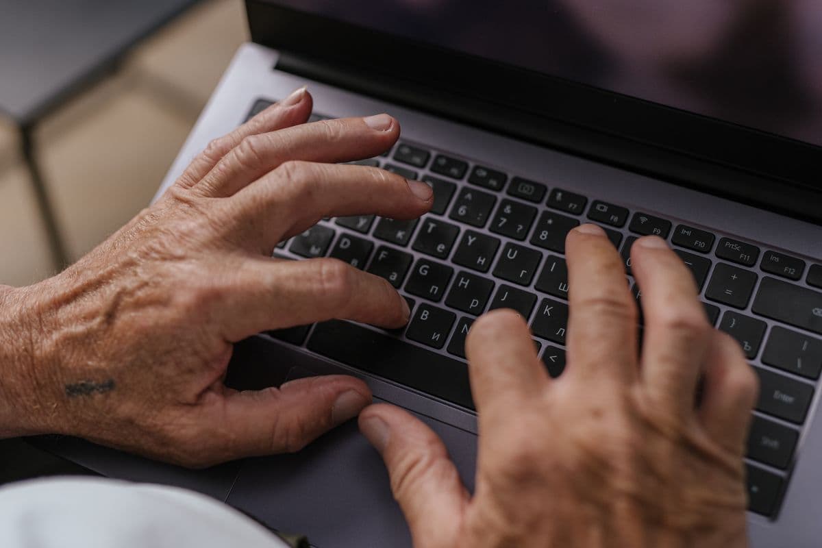 Older hands typing on a laptop keyboard with small, low-contrast key labels